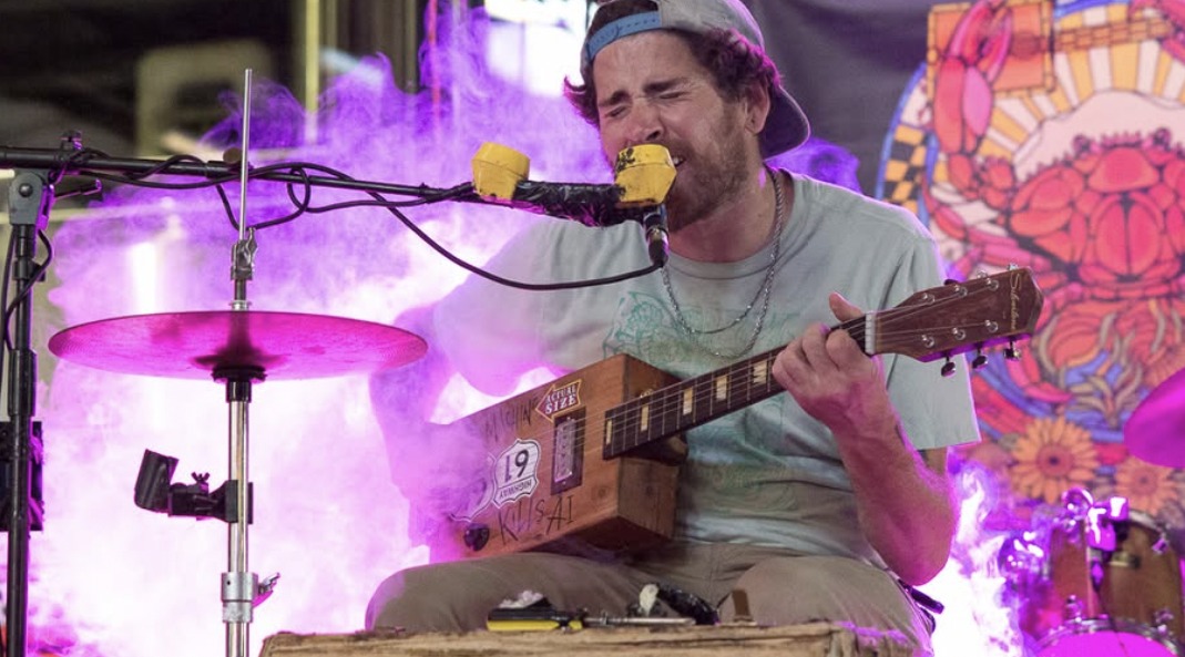 A performer in a backwards cap sings into a telephone-handset microphone while playing a cigar box guitar on a smoke-filled stage